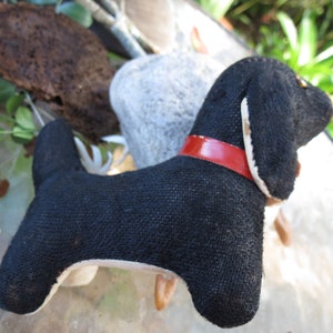 May include: A handmade black dog plush toy with a red collar. The dog has a long body and floppy ears. The toy is made of a textured fabric and has white accents on the ears and belly. The dog is a charming decorative item.