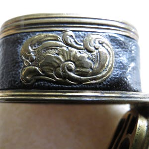 Op de afbeelding: Close-up van een zwarte lederen en gouden metalen decoratieve trim met een bloemmotief. De trim heeft een getextureerd oppervlak en een reli&euml;fdesign.