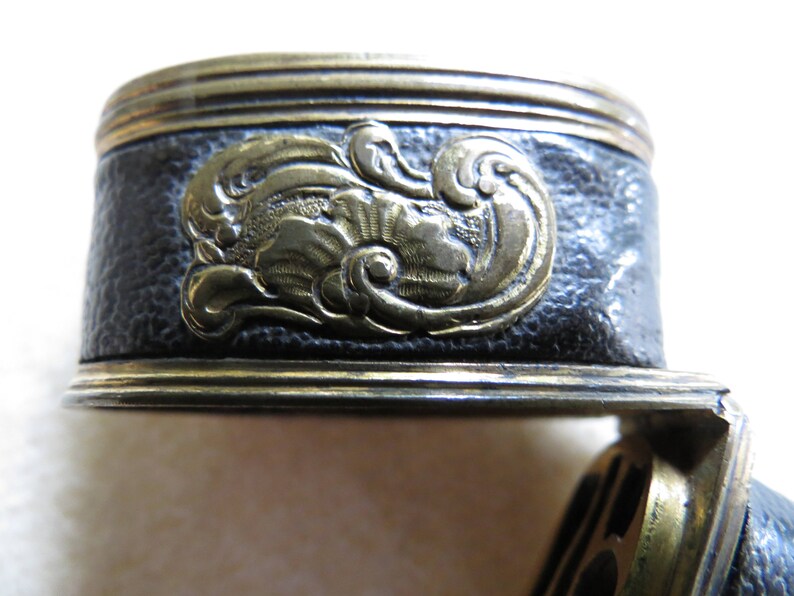 Op de afbeelding: Close-up van een zwarte lederen en gouden metalen decoratieve trim met een bloemmotief. De trim heeft een getextureerd oppervlak en een reli&euml;fdesign.