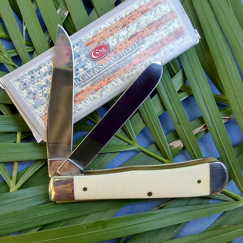 May include: A Case XX pocket knife with a white bone handle and a stainless steel blade. The knife is closed and the handle has two brass pins. The knife is resting on a bed of green leaves.