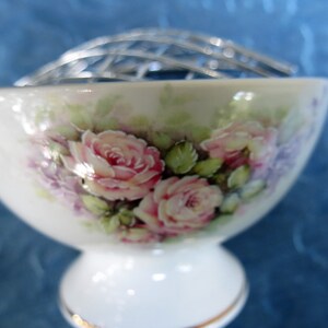 Puede incluir: Un florero de porcelana blanca con base y rejilla de metal plateado. El cuenco est&aacute; decorado con rosas rosas y flores moradas. El borde est&aacute; adornado con oro.