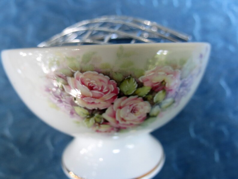 Puede incluir: Un florero de porcelana blanca con base y rejilla de metal plateado. El cuenco est&aacute; decorado con rosas rosas y flores moradas. El borde est&aacute; adornado con oro.