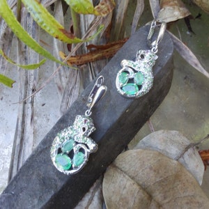 May include: A pair of silver earrings featuring a chameleon design with green gemstones. The earrings are set against a backdrop of brown wood and green leaves.