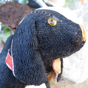 May include: A handmade black dog plush toy with a red collar. The dog has a yellow eye and tan nose and ears. The plush is made of a textured fabric and has stitched details.