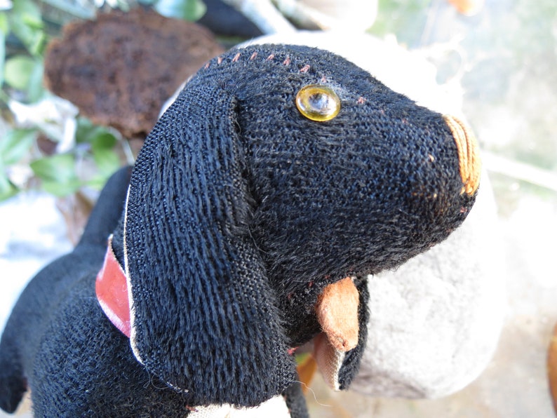May include: A handmade black dog plush toy with a red collar. The dog has a yellow eye and tan nose and ears. The plush is made of a textured fabric and has stitched details.