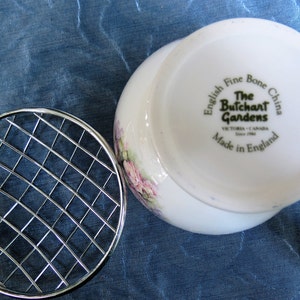 Puede incluir: Un jarr&oacute;n blanco de porcelana fina inglesa con un dise&ntilde;o floral y un salvaflores de metal plateado. El jarr&oacute;n tiene el texto "The Butchart Gardens, Victoria Canada, Made in England". El salvaflores tiene un patr&oacute;n de rejilla.