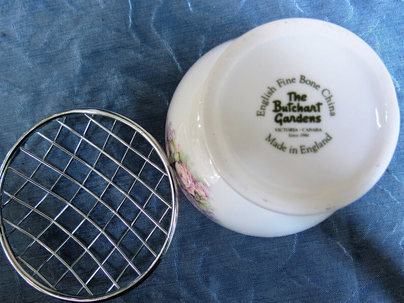 Puede incluir: Un jarr&oacute;n blanco de porcelana fina inglesa con un dise&ntilde;o floral y un salvaflores de metal plateado. El jarr&oacute;n tiene el texto "The Butchart Gardens, Victoria Canada, Made in England". El salvaflores tiene un patr&oacute;n de rejilla.