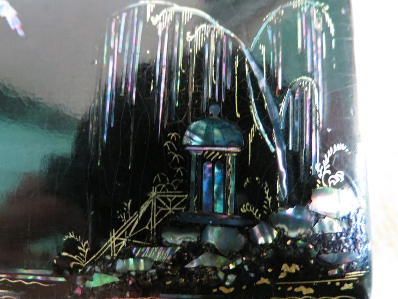 Pu&ograve; includere: Una scatola laccata nera con un design decorativo di un pagoda e alberi realizzati in madreperla iridescente. Il design &egrave; su uno sfondo nero.