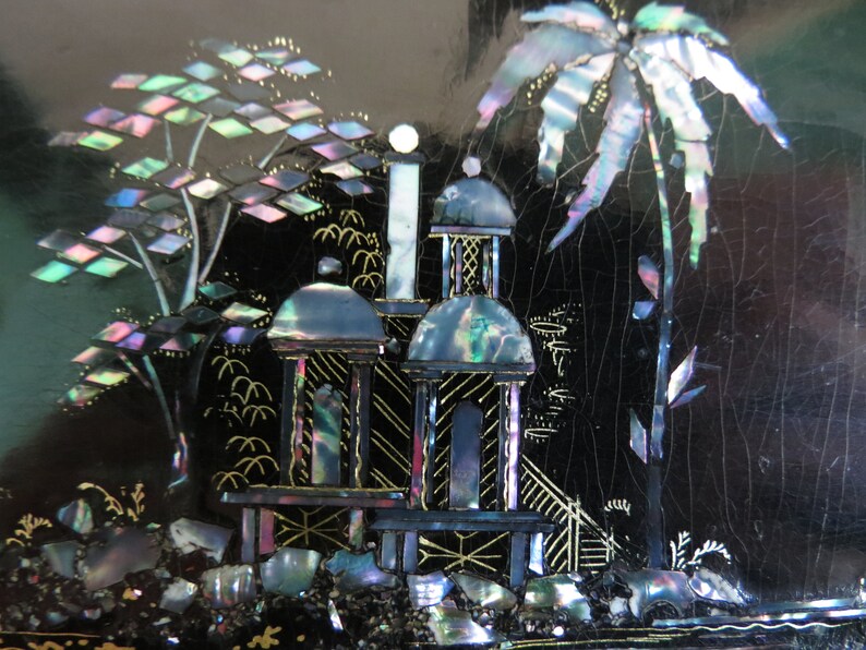 Pu&ograve; includere: Una scatola laccata nera con una scena decorativa di una palma e due strutture simili a pagode realizzate in madreperla iridescente. La scena &egrave; su uno sfondo nero con accenti dorati.