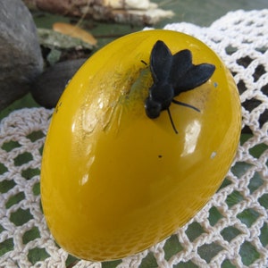 Pu&ograve; includere: Un uovo giallo con una mosca di plastica nera in cima. L'uovo &egrave; su una superficie di uncinetto bianca.