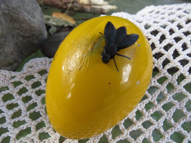 Pu&ograve; includere: Un uovo giallo con una mosca di plastica nera in cima. L'uovo &egrave; su una superficie di uncinetto bianca.