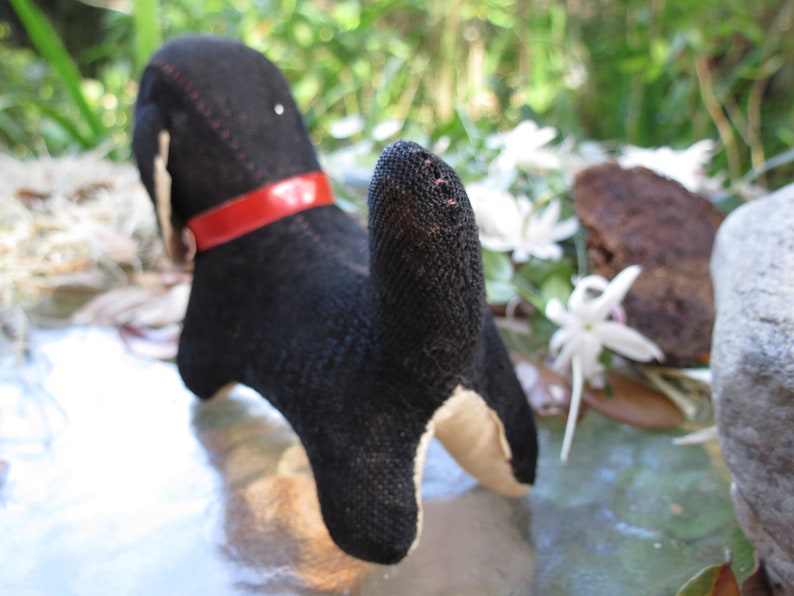 May include: A handmade black and tan dog plush toy with a red collar. The dog is made of fabric and has a stitched design. The toy is positioned on a reflective surface with a blurred background of greenery and white flowers.