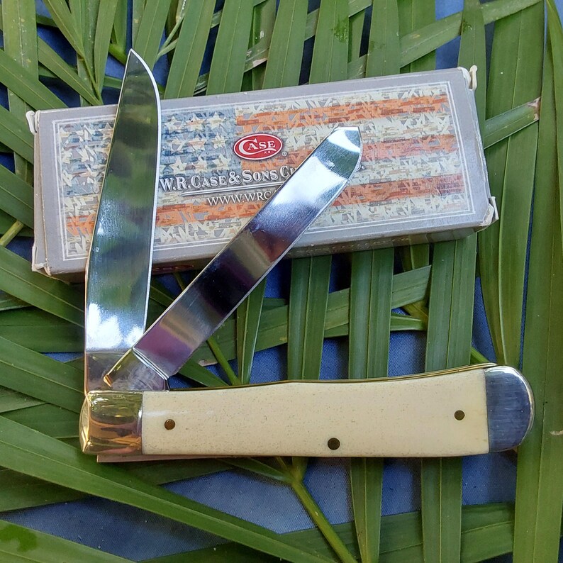 May include: A Case brand pocket knife with a white bone handle and a stainless steel blade. The knife is closed and the blade is visible. The knife is sitting on a green leafy background. The box for the knife is visible in the background.