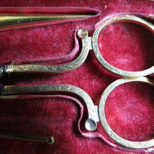 Pu&ograve; includere: Un set di forbici antiche color oro con disegni decorativi, riposanti in una scatola foderata in velluto rosso.