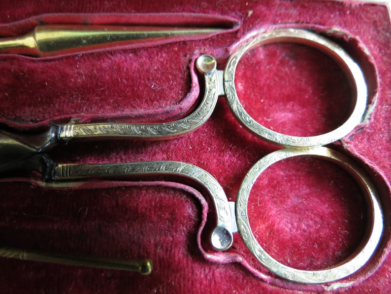 Pu&ograve; includere: Un set di forbici antiche color oro con disegni decorativi, riposanti in una scatola foderata in velluto rosso.