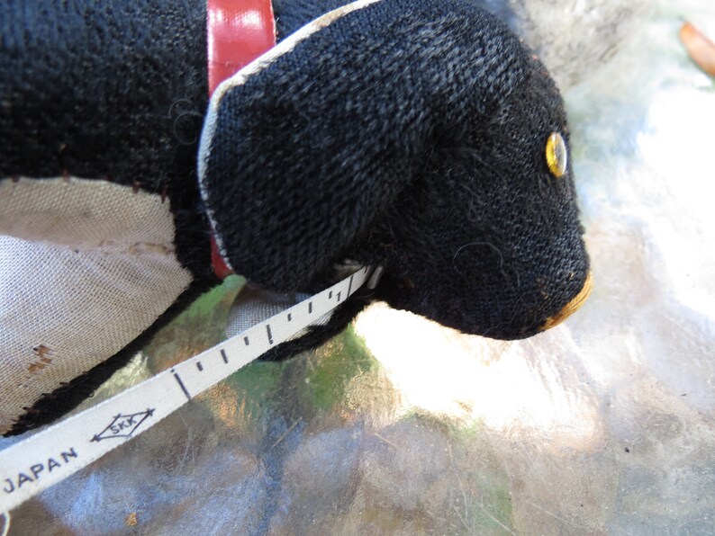 May include: A black stuffed dog with a red collar and a white and black measuring tape. The dog has a yellow eye and a tan nose. The measuring tape is marked in inches and has the word "JAPAN" printed on it.