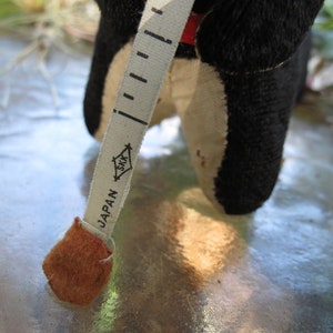 May include: A vintage black and cream-colored stuffed animal with a red accent, holding a measuring tape. The tape is marked with black increments and the word "JAPAN". The tape measure is beige with the logo "SKK".