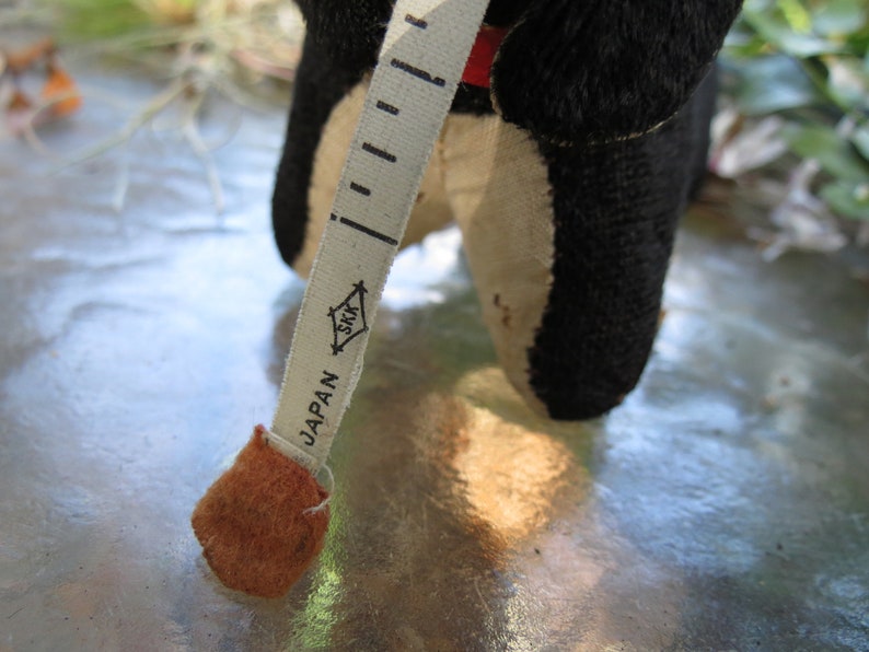 May include: A vintage black and cream-colored stuffed animal with a red accent, holding a measuring tape. The tape is marked with black increments and the word "JAPAN". The tape measure is beige with the logo "SKK".