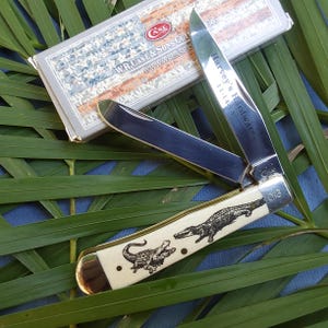 May include: A Case XX pocket knife with a white bone handle featuring an alligator design. The blade is open and the knife is engraved with "Harvey's Hardware 11-1-14".