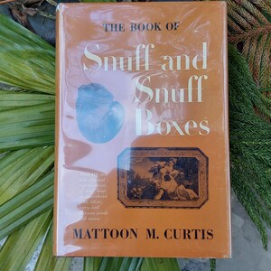 May include: A hardcover book titled "THE BOOK OF Snuff and Snuff Boxes" with an orange cover. The cover features a blue illustration of a snuff box and a framed image. The author's name, MATTOON M. CURTIS, is printed at the bottom.