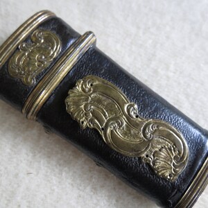 Op de afbeelding: Een zwarte leren schede met gouden metalen accenten en sierlijke ontwerpen. De schede heeft een decoratief, reli&euml;fpatroon van gestileerde golven.