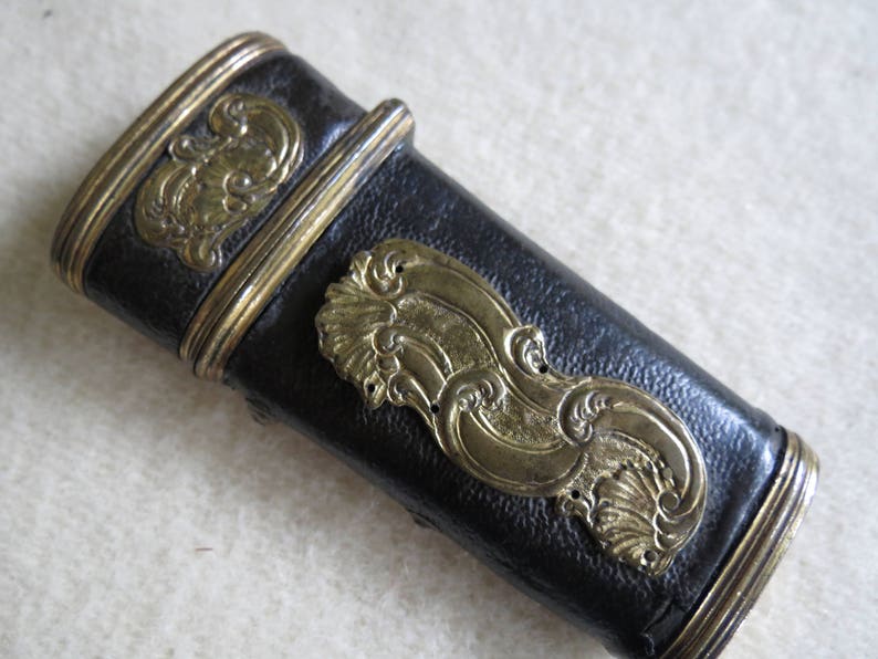 Op de afbeelding: Een zwarte leren schede met gouden metalen accenten en sierlijke ontwerpen. De schede heeft een decoratief, reli&euml;fpatroon van gestileerde golven.