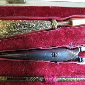 Pu&ograve; includere: Un kit da cucito vintage con un infila-ago dorato, un paio di piccole forbici argentate e un ago dorato con un manico decorativo. Gli strumenti sono esposti in una scatola foderata in velluto rosso.