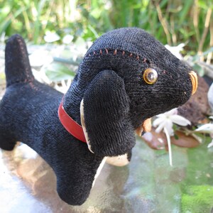 May include: A handmade black dog plush toy with a red collar and stitched details. The dog has large, yellow eyes and white accents on its paws and ears. The toy is made of a textured fabric.