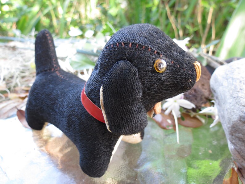 May include: A handmade black dog plush toy with a red collar and stitched details. The dog has large, yellow eyes and white accents on its paws and ears. The toy is made of a textured fabric.