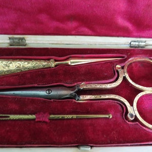 May include: A set of antique gold-toned sewing tools in a red velvet-lined wooden box. The set includes a pair of scissors, a needle threader, and a bodkin.