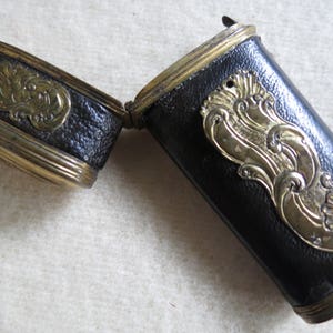 Op de afbeelding: Een zwart leren etui met gouden accenten en sierlijke ontwerpen. Het etui is scharnierend en opent om een leeg interieur te onthullen.