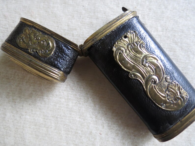 Op de afbeelding: Een zwart leren etui met gouden accenten en sierlijke ontwerpen. Het etui is scharnierend en opent om een leeg interieur te onthullen.