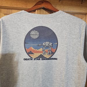 Peut inclure: T-shirt gris chiné avec un motif circulaire représentant l'Étoile de la Mort, un marcheur AT-AT et un paysage sous un ciel étoilé. Le texte "DEATH STAR ENGINEERING" est imprimé en dessous.