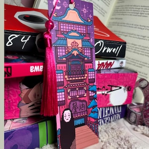 May include: A rectangular bookmark featuring pixel art of a multi-story building with blue roofs and pink walls, a black-robed figure with a white mask at the base, and a bright pink tassel attached to the top through a small circular hole.