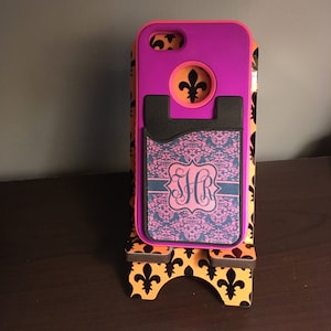 May include: A purple and pink phone case with a black card holder. The card holder has a pink and black damask pattern with the monogram "SJR" in the center. The case is on a stand with a fleur-de-lis design.