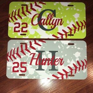 May include: Two rectangular license plate covers featuring a baseball stitch pattern in red. The top plate is lime green with the name Callyn and number 22. The bottom plate is light blue with the name Hunter and number 25. Both have mounting slots.