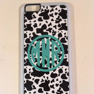 May include: A white phone case with a black and white cow print design. The case has a teal monogram in a circle that says "MMR".
