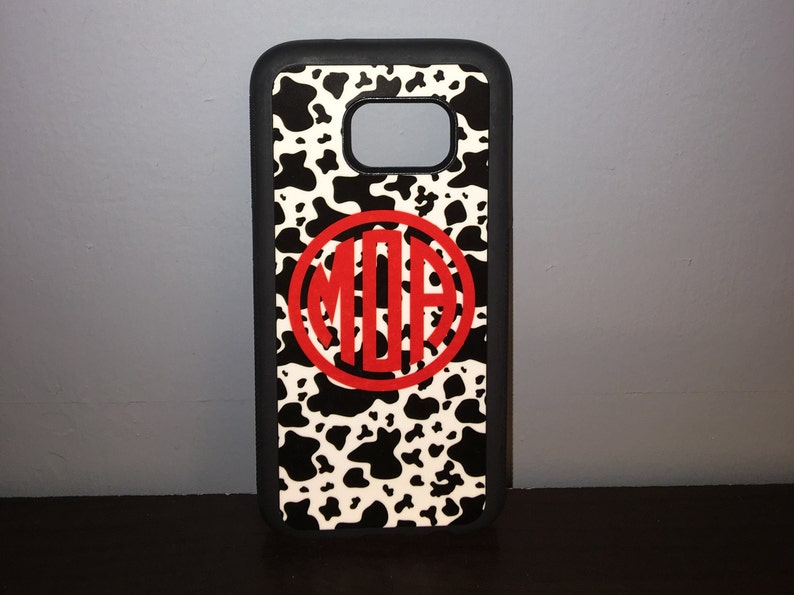 May include: A black phone case with a white and black cow print design. A red circle with the letters "MOA" in white is centered on the case.