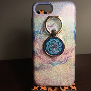 May include: A white glitter phone case with a blue and silver mandala design. The case has a ring holder with a blue and silver monogram that says "SVM".