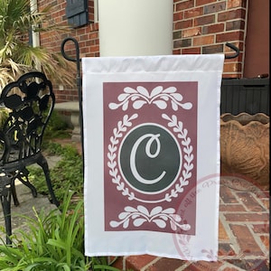 May include: A white garden flag with a maroon background featuring a white letter C inside a black circle surrounded by a wreath of white leaves. The wreath is on a maroon background.