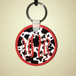 May include: A black and white cow print keychain with red trim and the letters "MR" in a white script font.