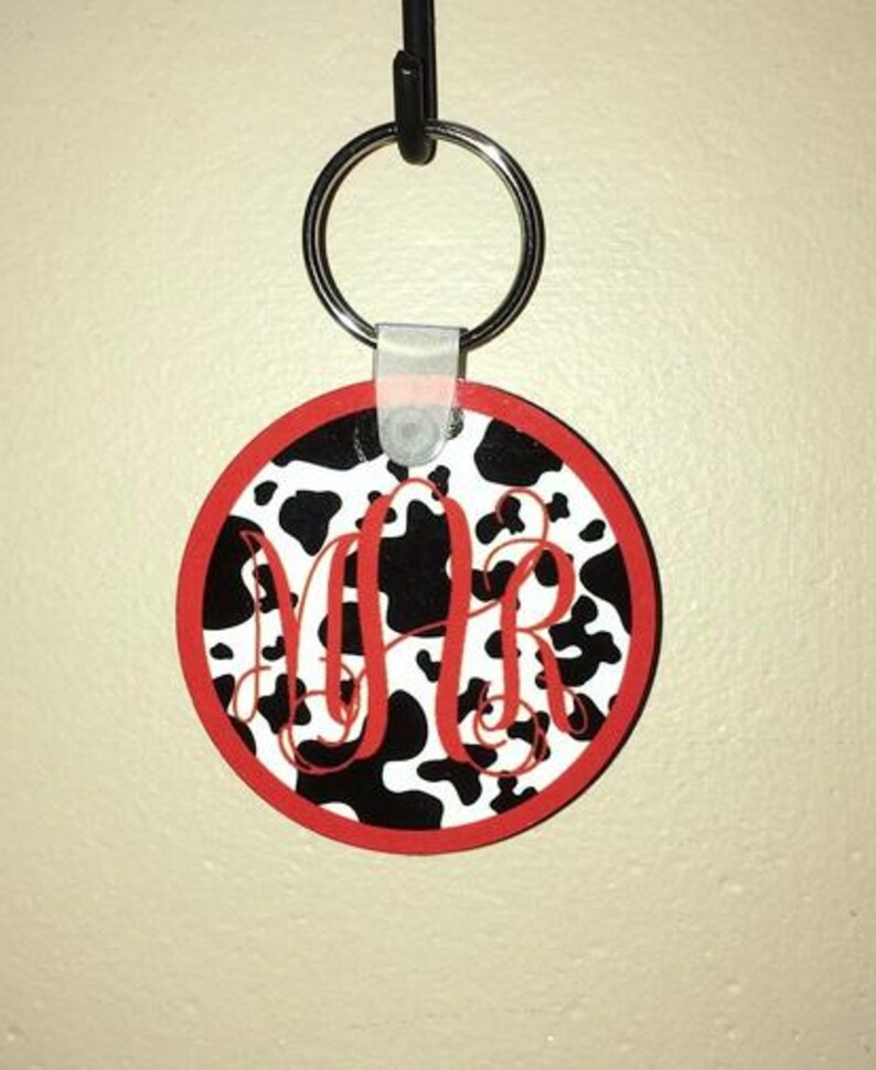 May include: A black and white cow print keychain with red trim and the letters "MR" in a white script font.