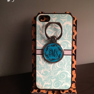 May include: A turquoise and white phone case with a swirl design. The case has a silver ring holder with a monogrammed circle in the center. The monogram is "BMN". The case is on a stand with a black and orange fleur-de-lis design.