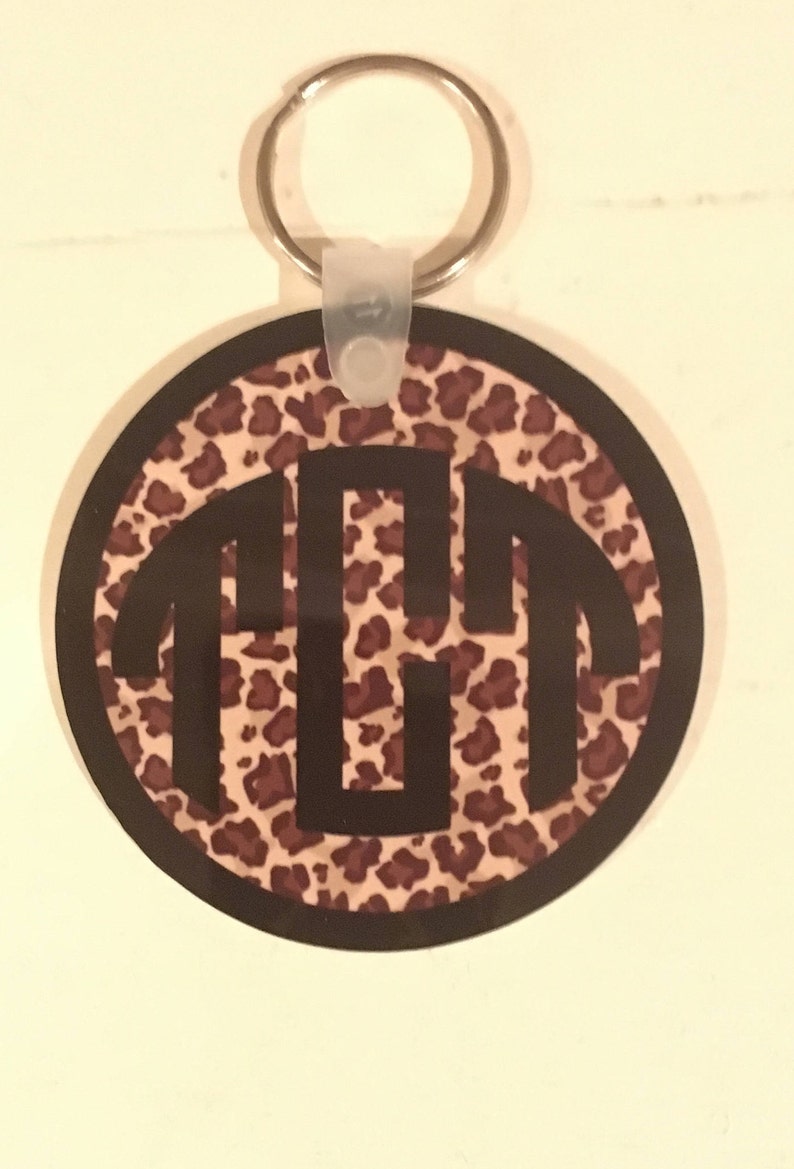 May include: A round keychain with a black and brown leopard print background. The keychain has a black monogram of the letters "TGT" in the center.