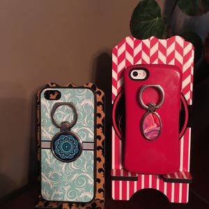 May include: Two phone stands with decorative patterns. The stands are made of wood and have a slot to hold a phone. The phone stands are decorated with a chevron pattern and a floral pattern. Each phone stand has a phone with a ring holder attached.