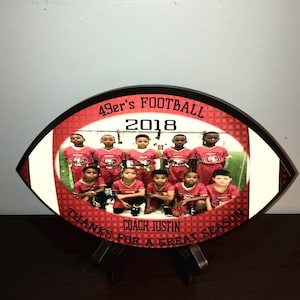 Może przedstawiać: Czerwono-biała tabliczka w kształcie piłki futbolowej ze zdjęciem młodzieżowej drużyny futbolowej w czerwono-złotych strojach. Na tabliczce widnieje tekst "49er's Football 2018" i "Dzięki za świetny sezon Trenerze Justin".