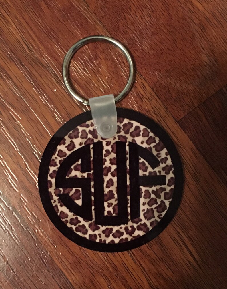 May include: A round keychain with a black and white leopard print background and a black monogram "SUF" in the center. The keychain has a silver metal ring.