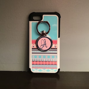 May include: A white phone case with a blue and pink geometric pattern. The case has a silver ring holder with a pink circle that says "A" in blue lettering.