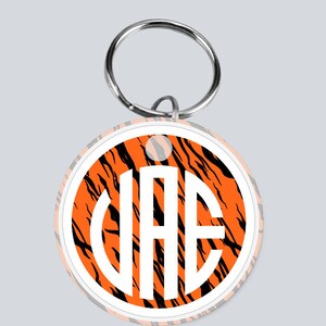 May include: A round orange and black tiger print keychain with a white circle in the center. The white circle has the letters "VAE" in black.