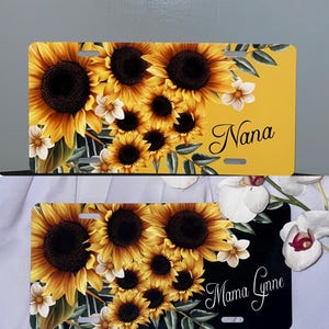 May include: License plates featuring a vibrant sunflower design. One plate has a yellow background with the word "Nana" in script, while the other has a black background with "Mama Lynne". Both plates are adorned with sunflowers and white flowers.